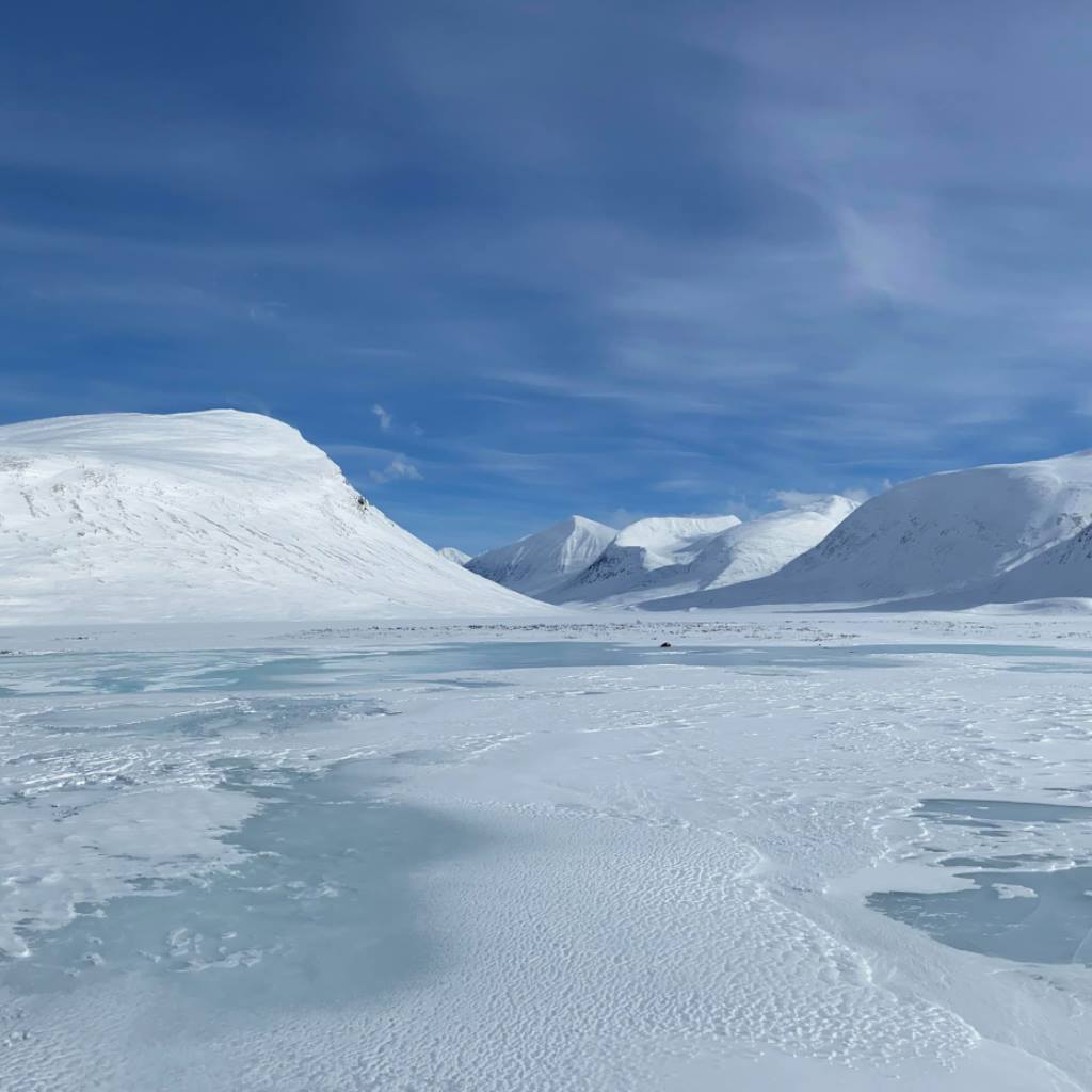 Dalen Alggavarre med grön nyis ovanpå snön på sjön Alggejavrre. Kapellet Alkavare ligger vid sjön och vid fjället Alggavarre.