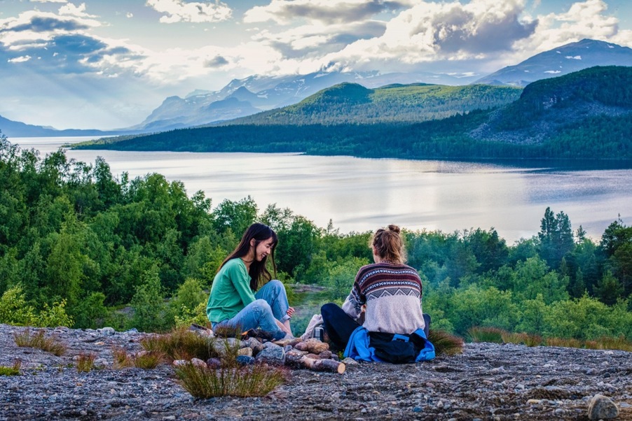 Två unga tjejer sitter vid en brasa med utsikt över fjället. 