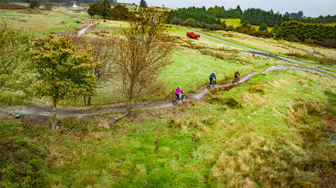 Flygbild över landskap med gräsängar och träddungar. Två personer syns, som cyklar mountainbike på en smal stig.