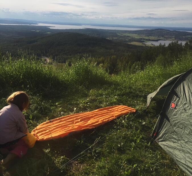 Foto: Victor Solli Ett barn blåser upp ett orange liggerunderlag brevid ett grönt tält, på en höjd. I bakgrunden syns utsikt över berg och sjöar.