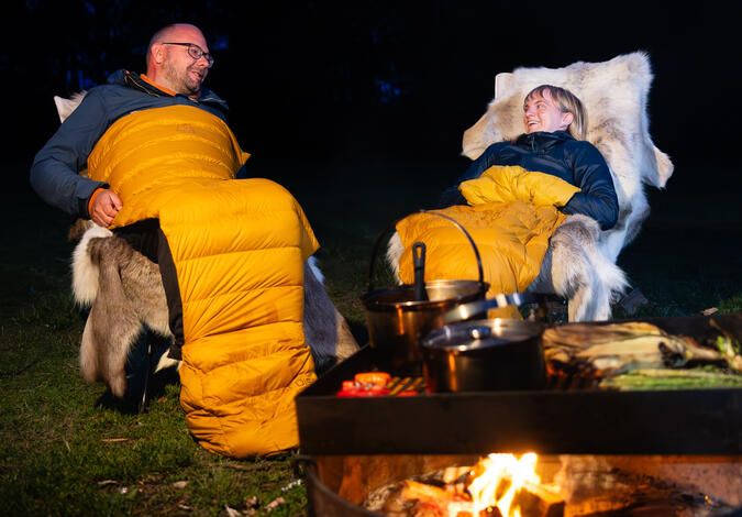 Foto: Piotr Två personer sitter framför en lägereld. Båda har den sovsäcksförstärkande produkten "Boost +8" från Nordisk lindade runt kroppen.