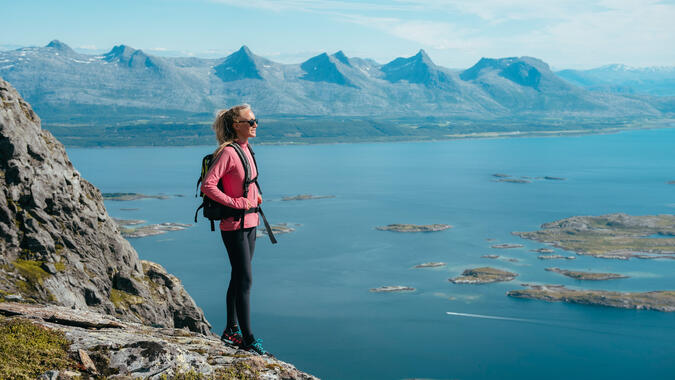 Utsikt över "De sju systrarna" från berget Dønnamannen. Foto: Terje Rakke, Nordic Life / Visit Helgeland En kvinna står på ett berg, med utsikt över havet och bergskedjan De sju systrarna i Norge.