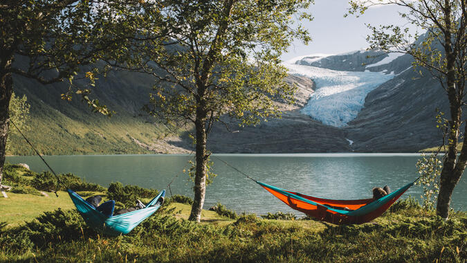 Koppla av i hängmatta med utsikt över glaciären Svartisen vid Engabrevatnet. Foto: Emilie Solbakken / Visit Helgeland Två personer ligger i hängmattor med utsikt över glaciären Svartisen.