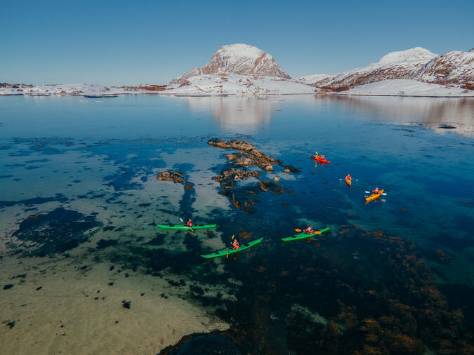 Vinterpaddling vid Støtt i Meløy. Foto: Emilie Solbakken / Visit Helgeland Sex personer paddlar kajak på havet. I bakgrunden syns snöiga berg.