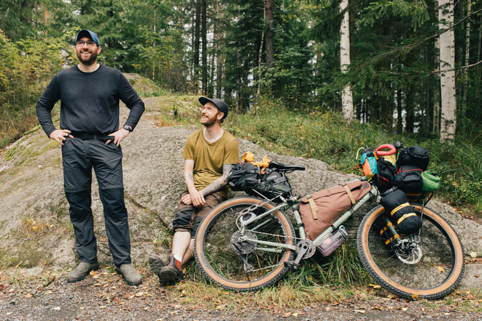 En man står till vänster, medan en annan man sitter lutad mot en sten mot vilken en fullpackad gravelbike vilar.