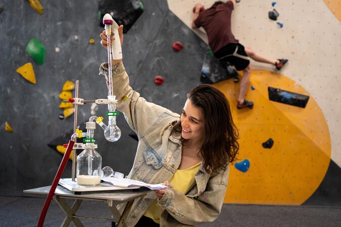 Dr. Anya Sherman samlar in partiklar från luften i Wiens boulderinghallar med en impinger. Foto: Aaron Kintzi / CeMESS En kvinna sitter och arbetar med provrör. I bakgrunden syns en klättrare på en klättervägg.