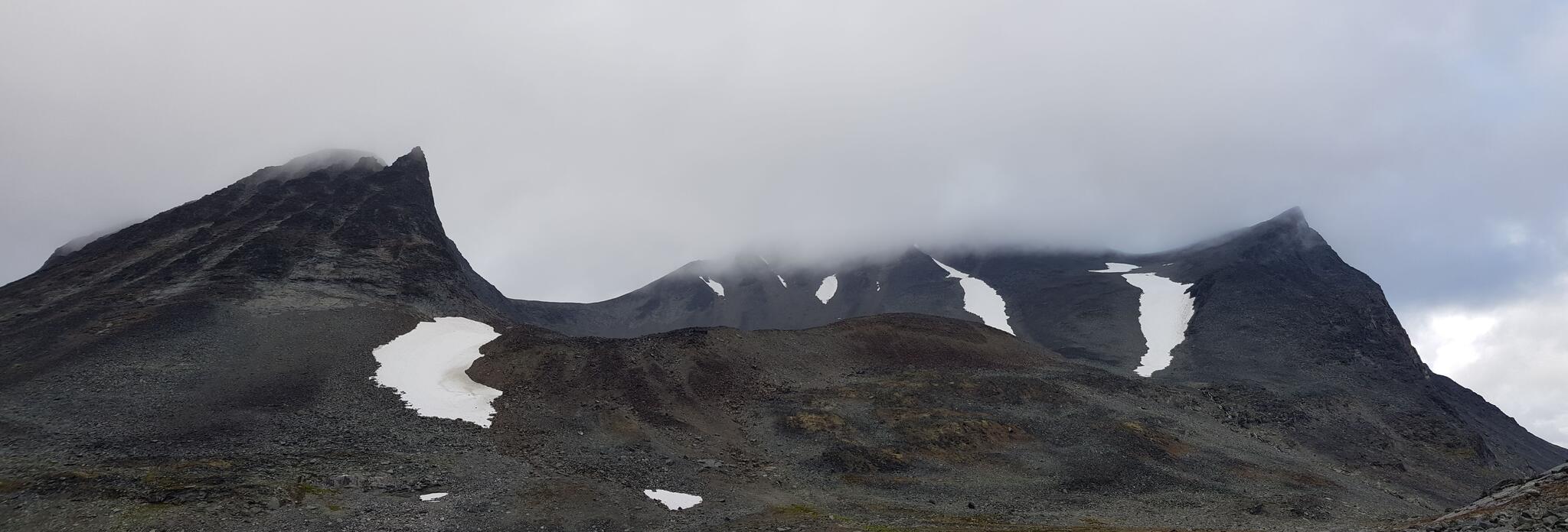 Šielmmáčohkkamassivet sett från Nallús västliga utlöpare. Längst till höger ses Kugghjulskammens västligaste topp. Šielmmáčohkkas stortopp är dold bland moln. Vi vandrade upp längs den vänstra kanten tillhörande snöfältet längst till höger (öster). Varnar för det något rödaktiga partiet i mitten av bilden - brant och risk för ras.
